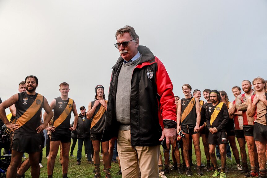 Man wearing black and red jacket stands on footy field with players behind him.