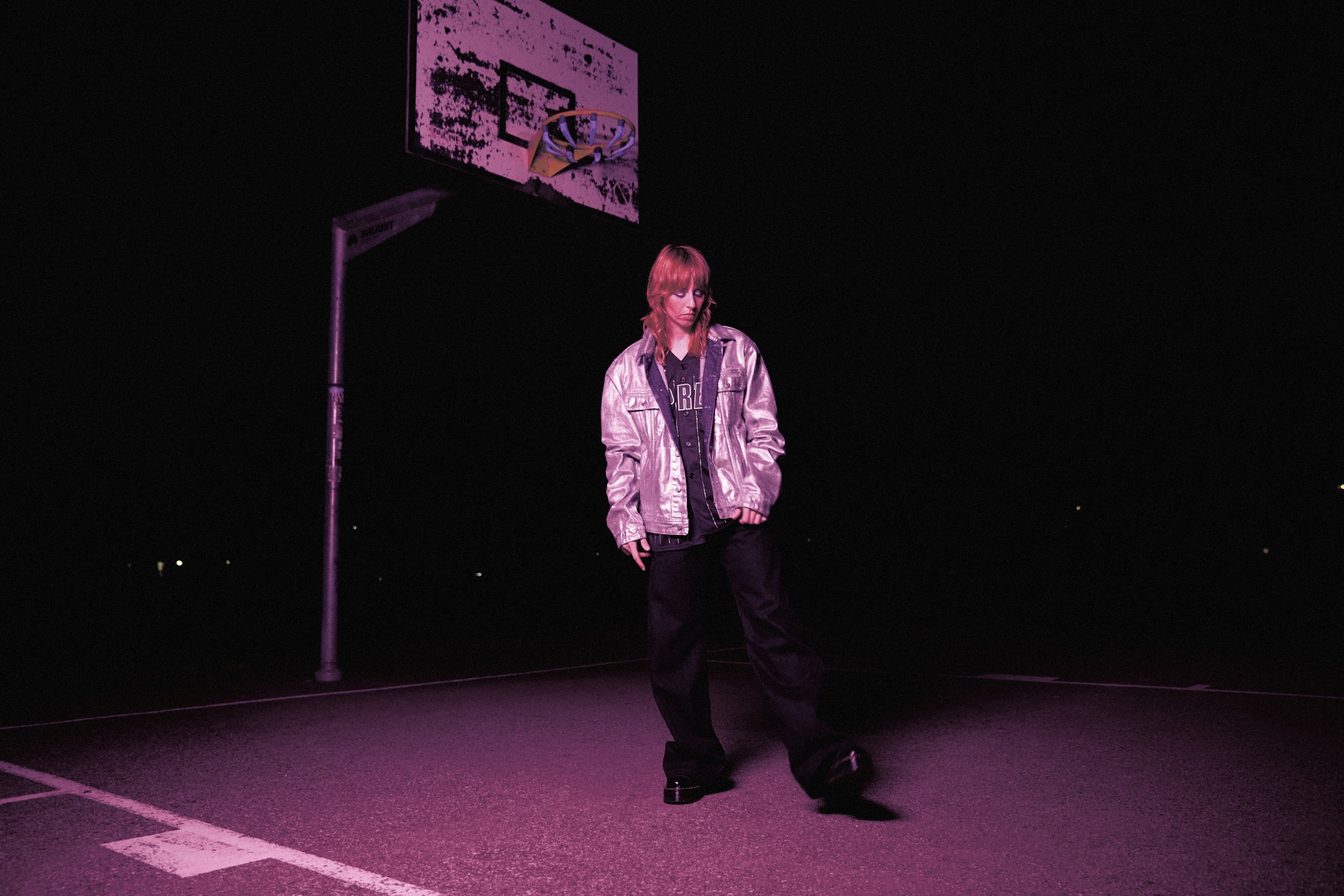 woman stands on basketball court at night wearing baggy clothes. a hoop hangs behind her