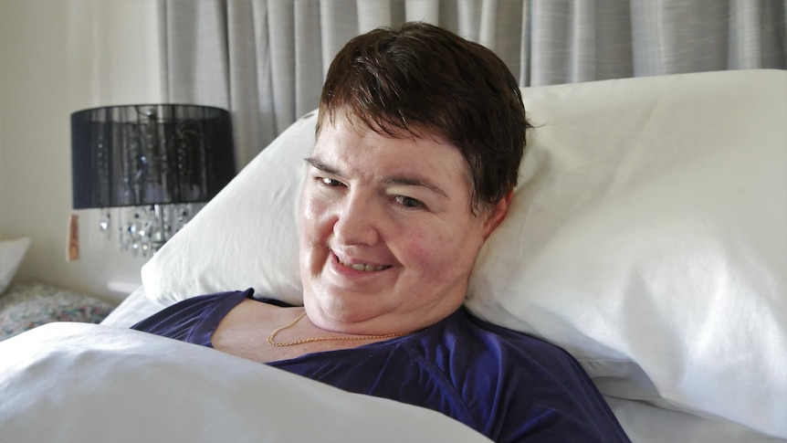 A woman lying in bed smiles. She is wearing a purple shirt and necklace. She has dark hair.