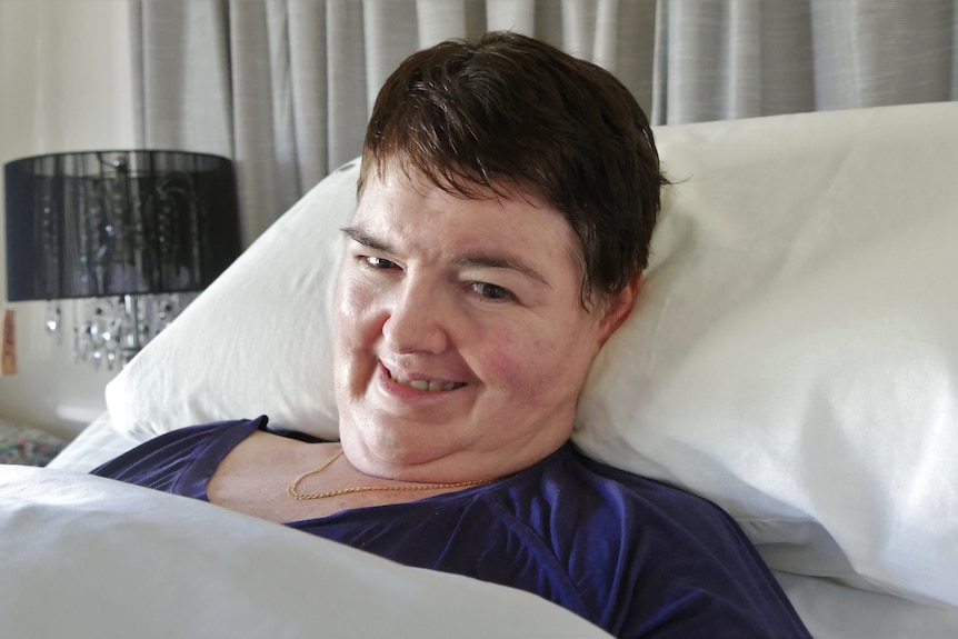 A woman lying in bed smiles. She is wearing a purple shirt and necklace. She has dark hair.