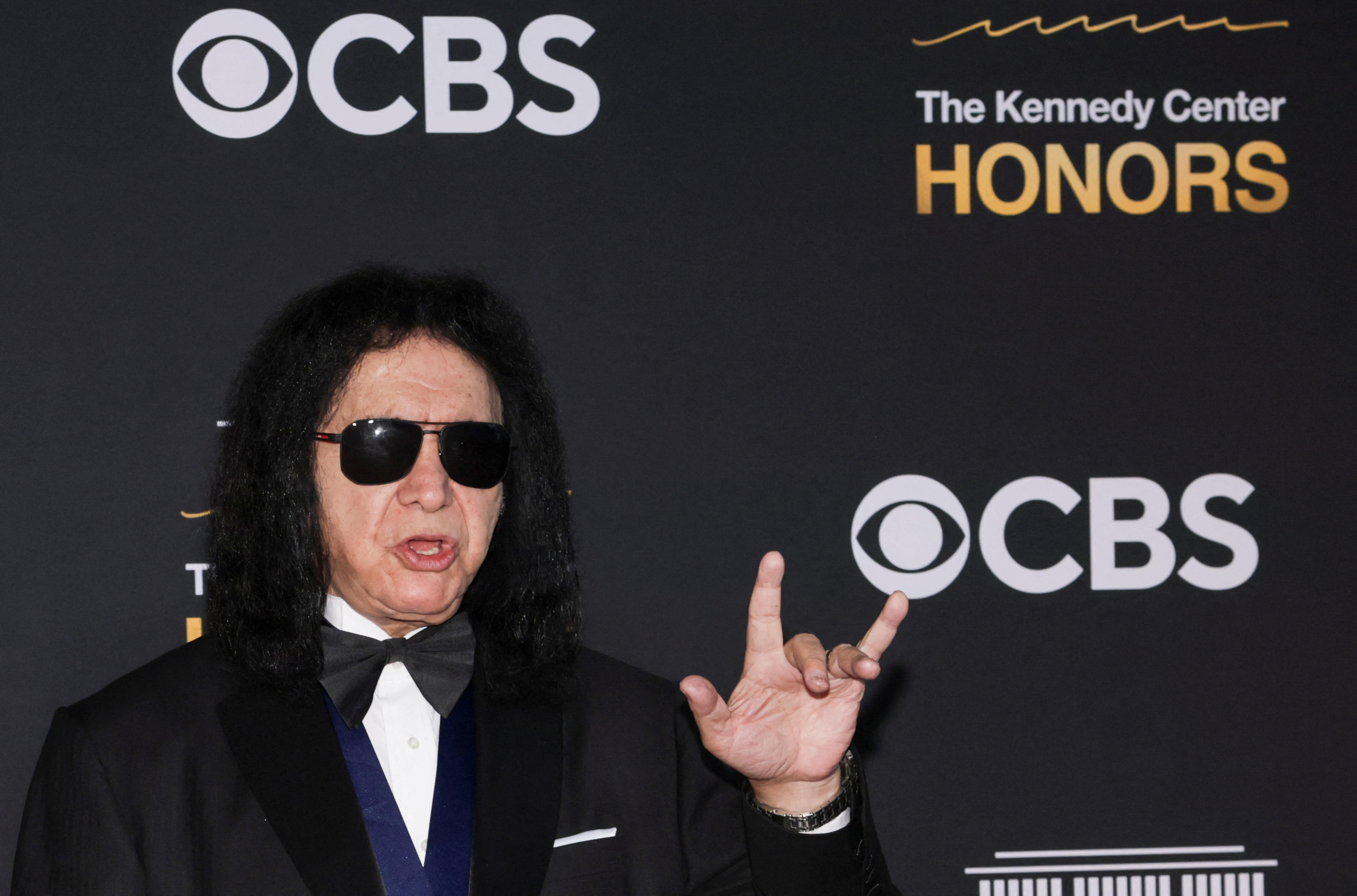Gene Simmons levanta una mano en un símbolo de rock mientras usa gafas de sol y un esmoquin sobre un fondo oscuro.
