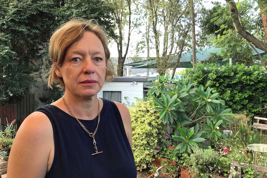 A woman stands in her backyard surrounded by plants and trees.