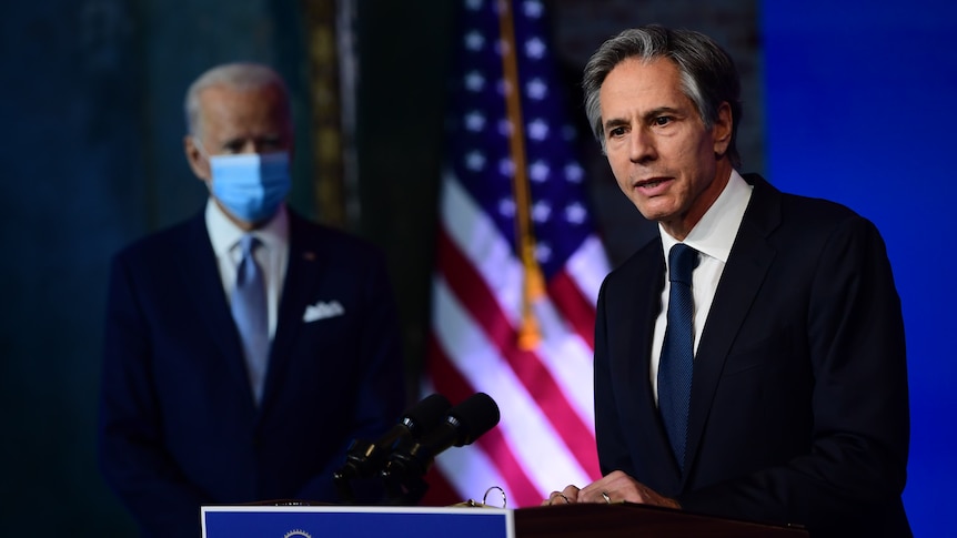 Antony Blinken stands behind a lecturn in mid-speech. Joe Biden stands wearing facemask, blurred in the background.