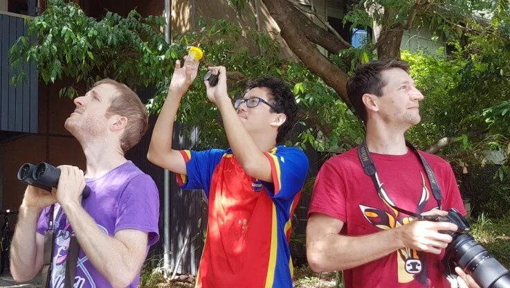Three young men standing outside holding binoculars and a camera