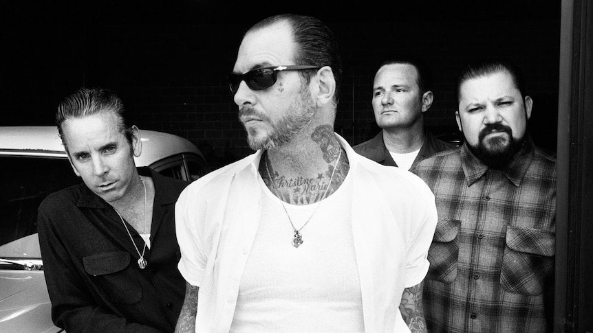 Black and white photo of four men from punk band Social Distortion standing beside an old car.,
