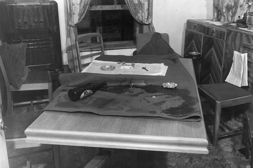 Black and white photo of a small dining room. Ashtray, knocked over beer bottle and cup on table. Dark stain on table cloth.