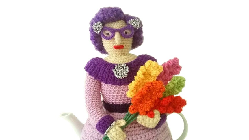 A crocheted Dame Edna holding a a bunch of gladioli.
