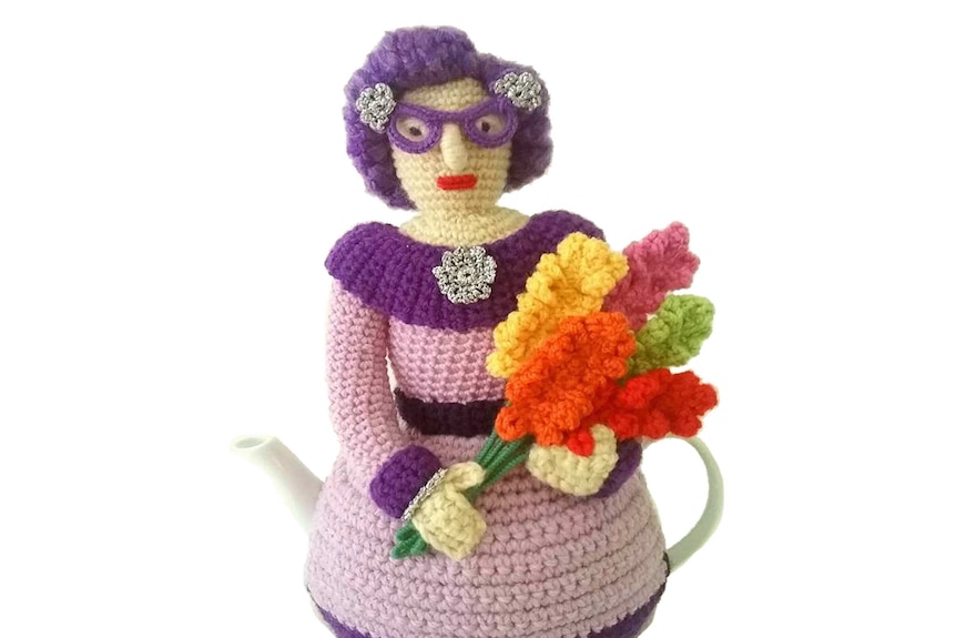 A crocheted Dame Edna holding a a bunch of gladioli.