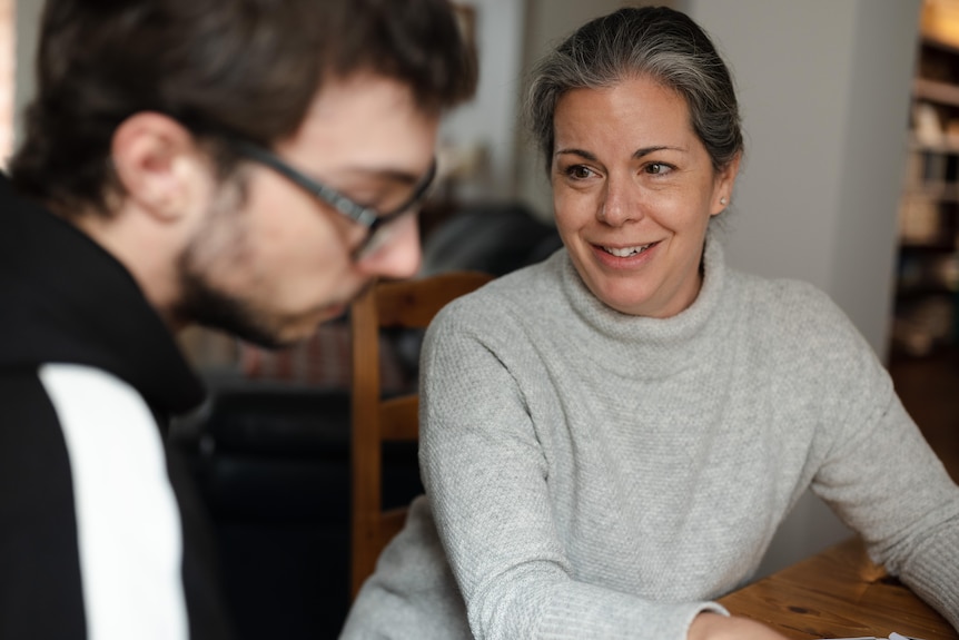 A woman smiles as she looks at the teenager sitting next to her at a table.