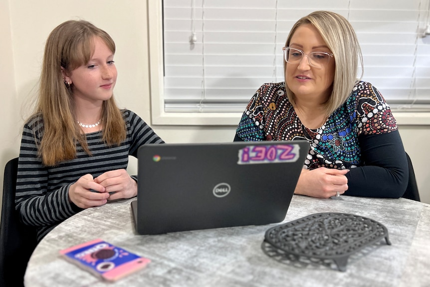 A girl and a woman sit at a round table indoors, looking at a laptop screen.