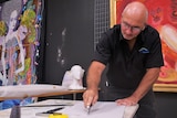 A bald man wearing a black shirt and spectacles, cuts a piece of cardboard with a knife, with colourful artworks behind.
