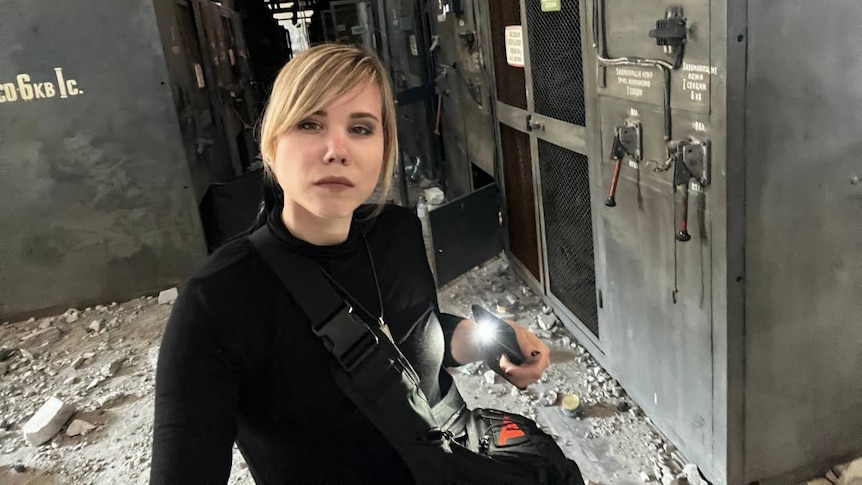 A young blonde woman dressed in black takes a selfie in a building surrounded by rubble