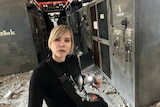 A young blonde woman dressed in black takes a selfie in a building surrounded by rubble