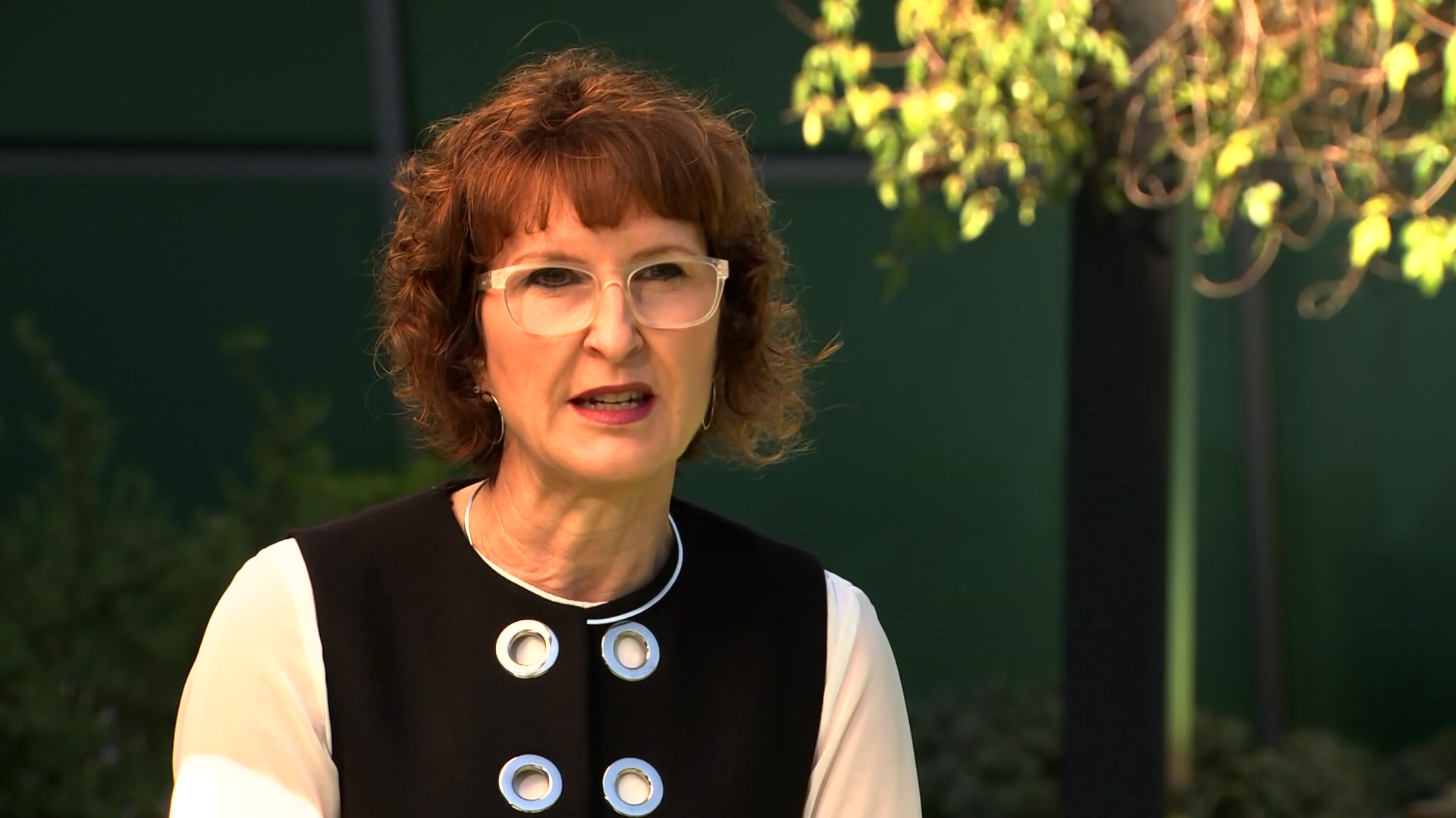 Woman in a black-and-white top talks to an interviewer out of frame. She has short orange hair and white eyeglasses.