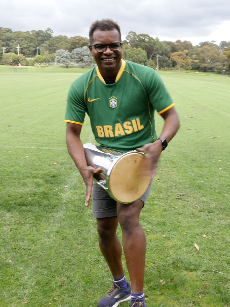 A man dressed in green shirt that reads 'Brasil' holds a drum and looks at the camera.