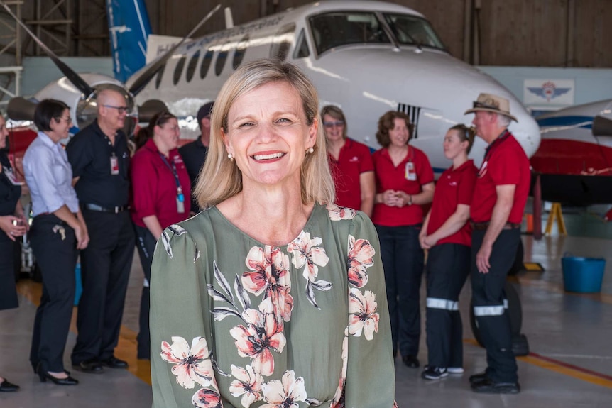 A woman with shoulder-length blonde hair smiles at the camera while a group of people stand in front of a plane behind her.