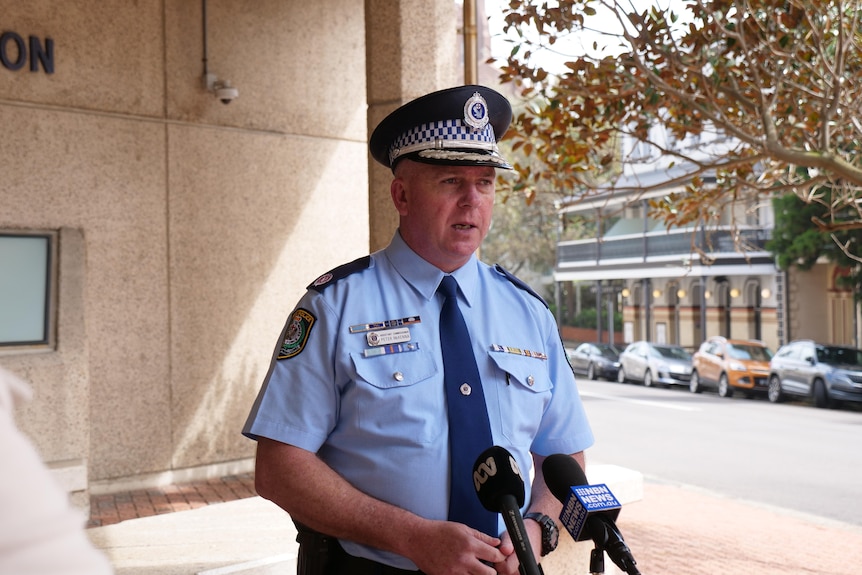 A uniformed police officer talks into two news microphones.