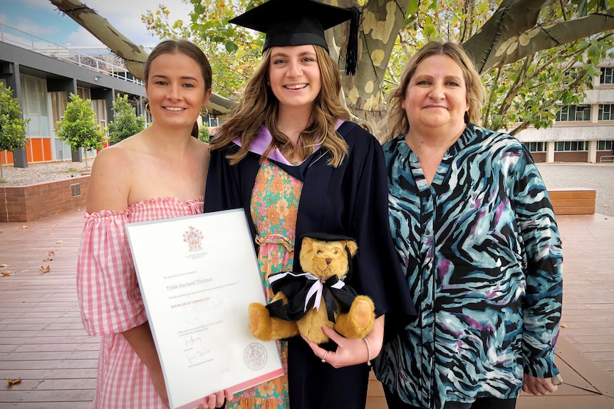 a lady stands in her graduation robes holding her degree and a teddy bear