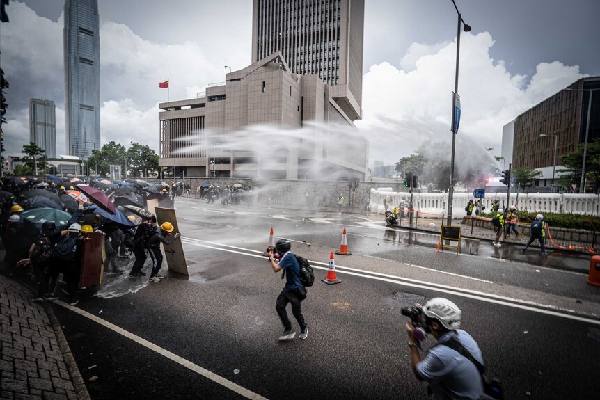 Protesters hold umbrellas and shields and brace themselves as a water cannon fires over head