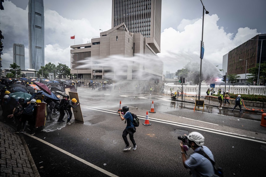 Protesters hold umbrellas and shields and brace themselves as a water cannon fires over head