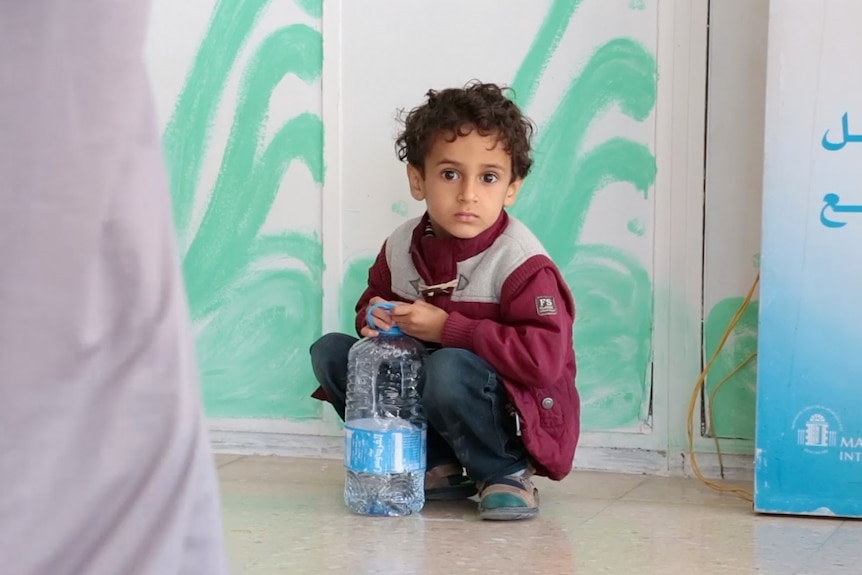 young frightened boy squats down in hospital corridor and looks at camera with wide eyes.