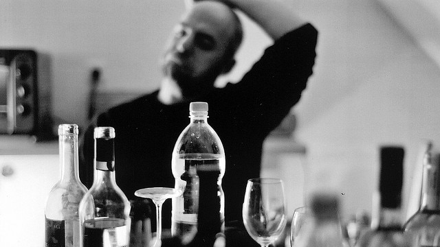 A black and white image of a man anxiously regarding several bottles of alcohol on a table.