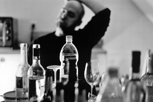 A black and white image of a man anxiously regarding several bottles of alcohol on a table.