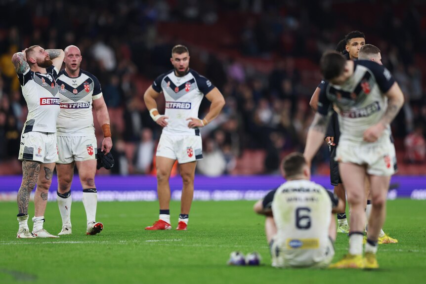 An England rugby league player clasps his hands behind his head and looks skywards as teammates look exhausted and dejected.