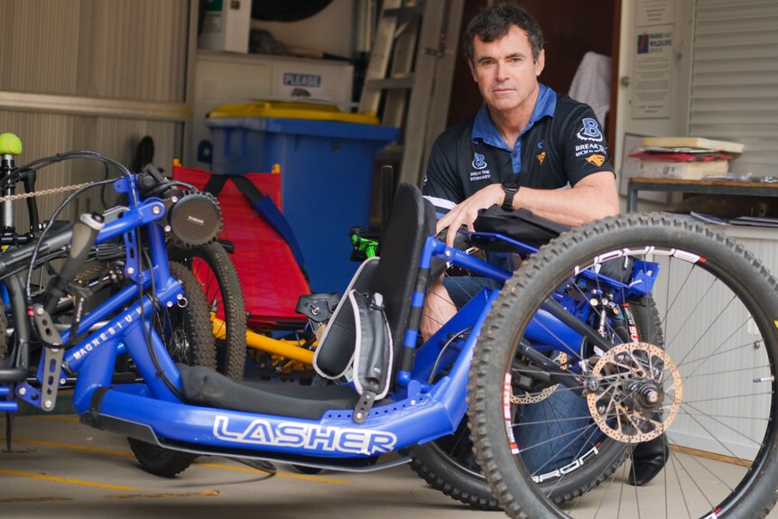A man crouched down next to an three-wheel adaptive mountain bike, looking at the camera.