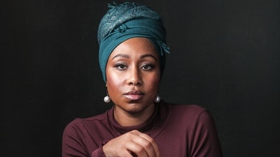 a woman wearing a blue headscarf looks directly into the camera, she's holding another scarf in her hand
