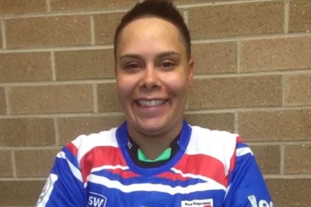 A woman smiles for the camera in front of a brick wall and is wearing a blue red and white rugby league guernsey.