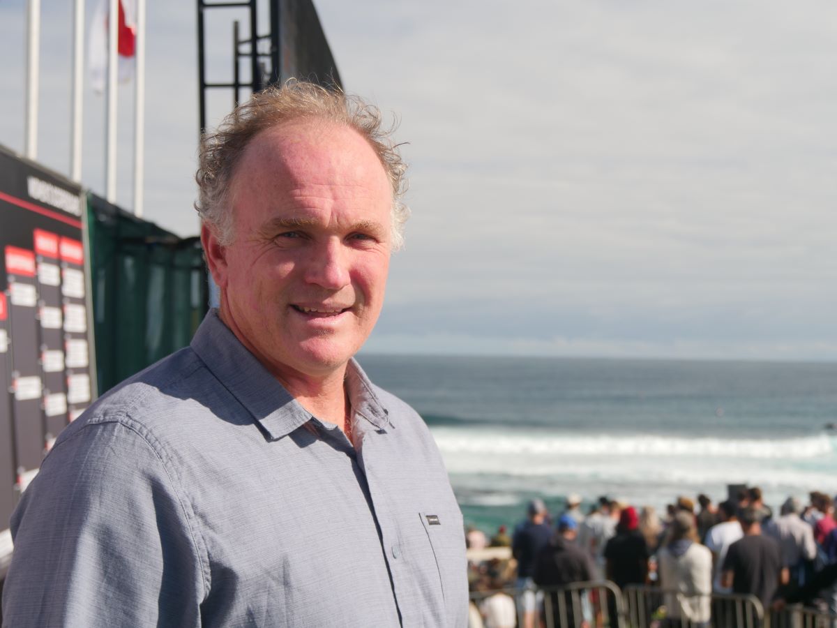 Andrew Stark stands near surf competition infrastructure with the ocean and waves in the background.