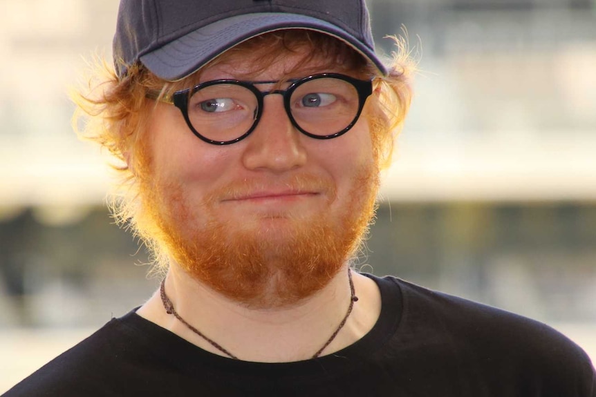 Musician Ed Sheeran wears glasses and a black cap as he smiles with Perth Stadium in the background.