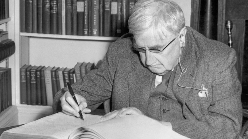 A black and white photo of an older Ralph Vaughan Williams sitting at a table and notating in a book.