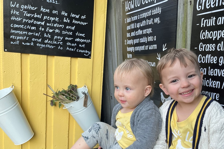 An image of Katie Irwin's children sitting on black composting bins infront of yellow hutch with chalk writing on blackboard