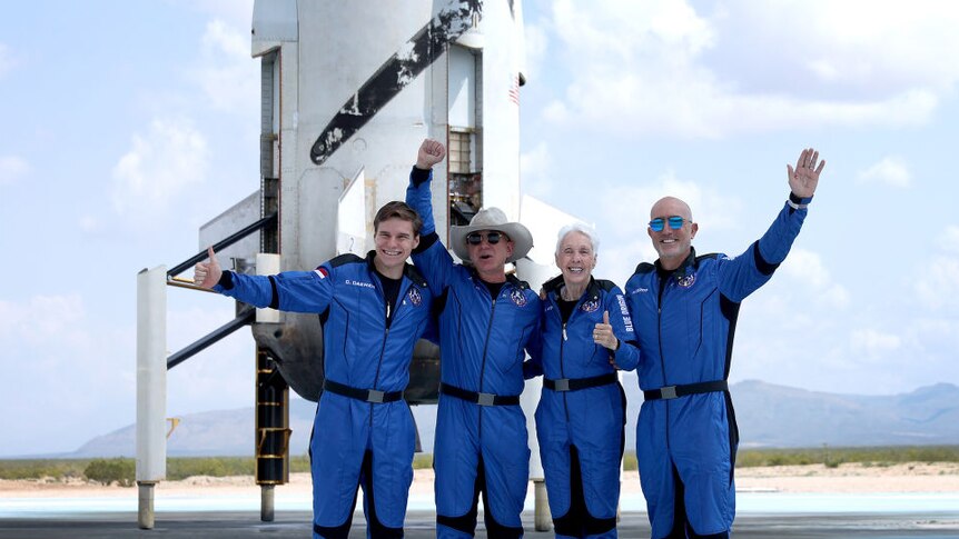 Jeff Bezos and three other space passengers in blue space uniforms grinning and posing in front of rocket