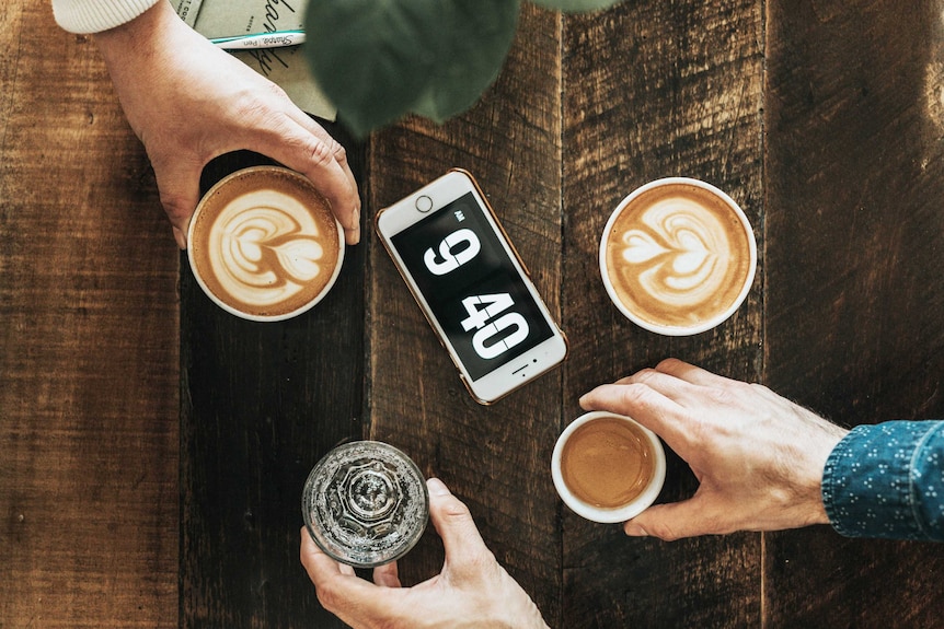 Aerial of people clutching cups of coffee around an digital clock on phone showing to represent timeliness in the workplace.