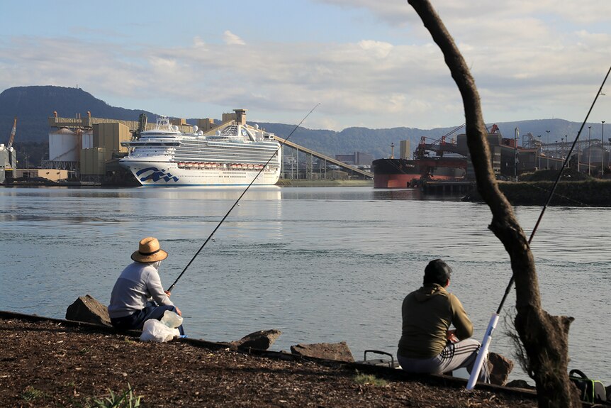 A pair of fishermen sit across the harbour from a cruise ship docked at an industrial port.