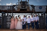 An informal bridal party stands in a rodeo bull ring underneath the cabin of a truck's prime mover.