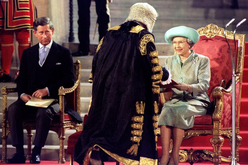 The Queen and Prince Charles sit in thrones while a man in a ceremonial clock and wig speaks to the Queen
