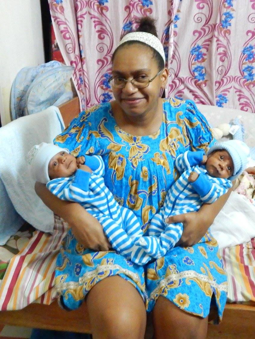 A woman wearing a blue dress sitting with her two baby sons dressed in matching blue striped suits. 