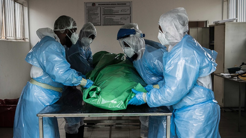 A group of people in full-body PPE lift a body wrapped in plastic off a metal table.