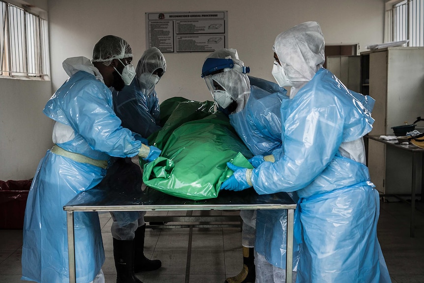 A group of people in full-body PPE lift a body wrapped in plastic off a metal table.