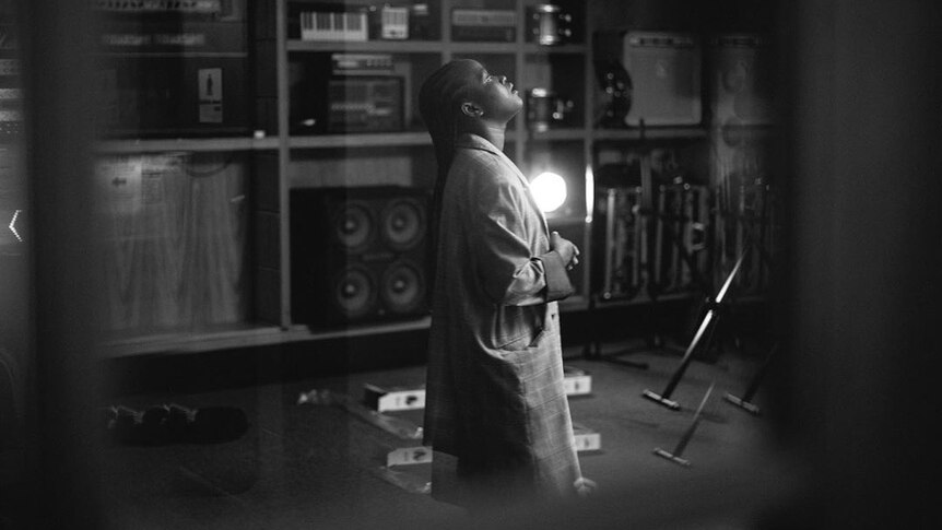 A young black woman stands in a room looking up at the ceiling with her hands clasped as if in prayer.