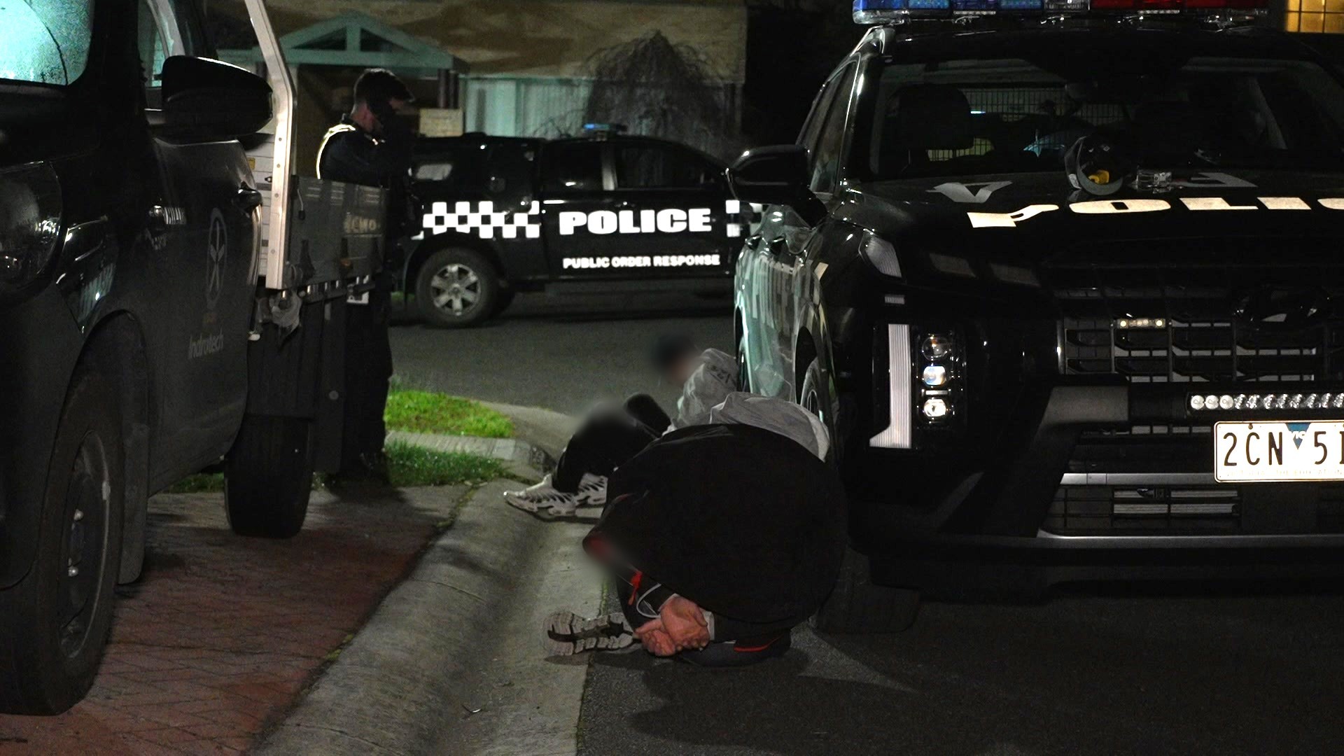 Two handcuffed people in grey and black clothing sit on the road against a black police car with their heads turned away.