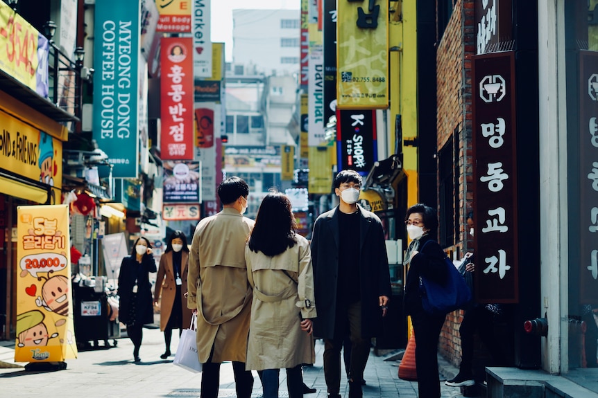 A man in a dark coat and white face mask walks through a busy shopping district in Seoul 