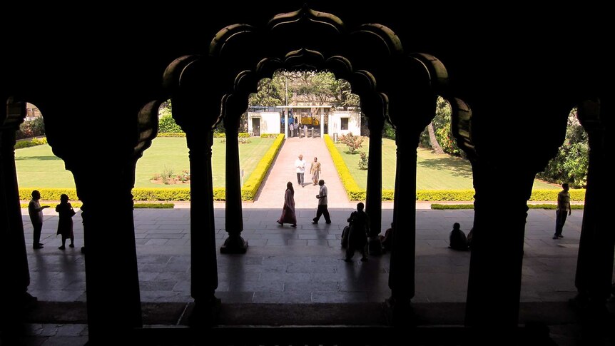 Looking out between dark, dimly lit columns in a palace to a path and entrance. People stand around in the sun.