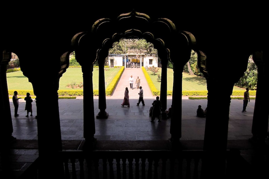 Looking out between dark, dimly lit columns in a palace to a path and entrance. People stand around in the sun.