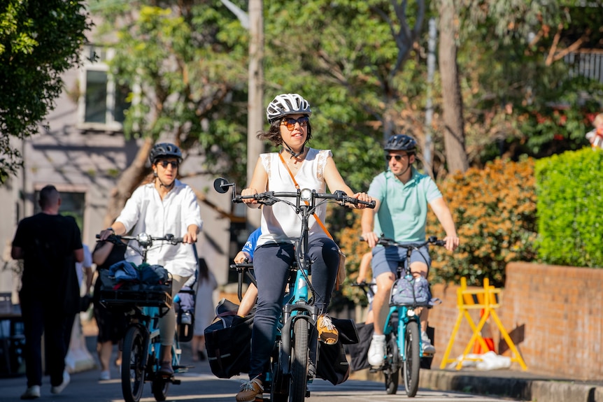 A woman in a white top rides a bike with a child sitting on the back. Two other cyclists ride behind.