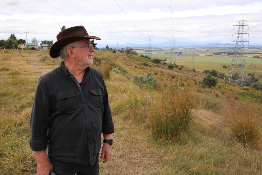 Man with a grey beard and big brimmed hat stands out in a field and smiles off into the distance